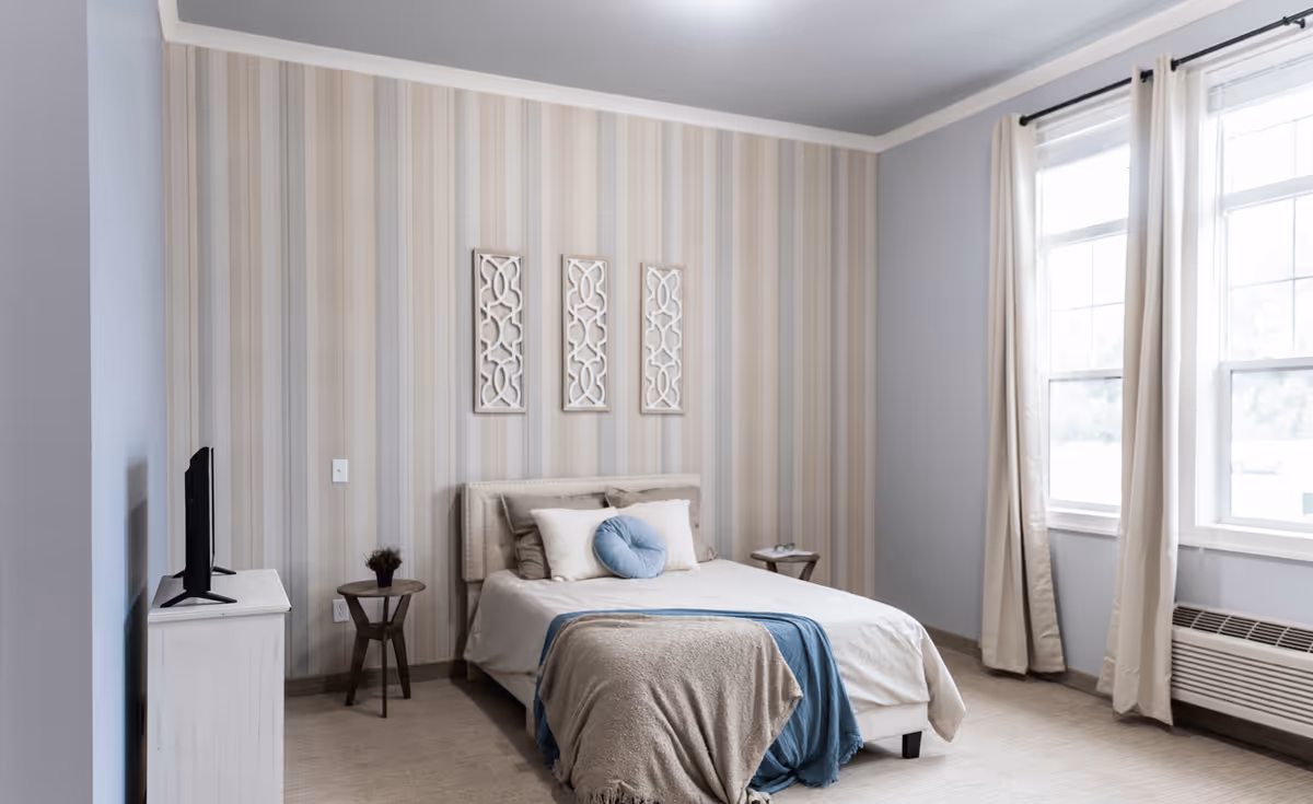 Bright, neatly decorated bedroom with a bed against a striped accent wall, windows with curtains, side tables and a TV on a dresser.