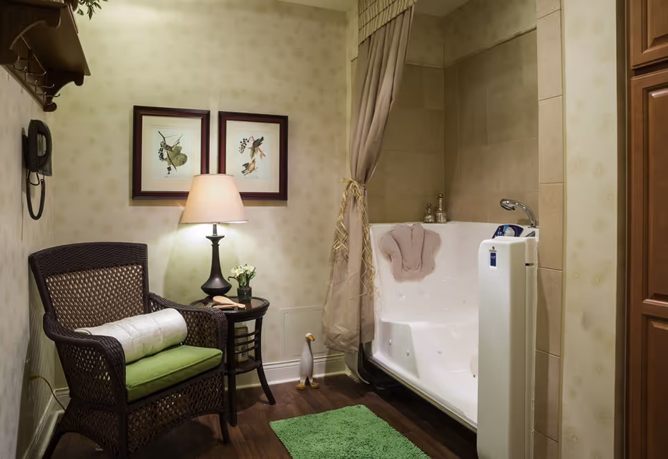 A cozy bathroom corner featuring a white walk-in bathtub with a beige towel draped over the backrest, a green bath mat on the wooden floor, and a wicker chair with a green cushion and white bolster pillow. Next to the chair is a small round table with a lamp, a small plant, and a brush. Two framed botanical prints hang on the wallpapered wall, and a small decorative duck figurine stands on the floor near the bathtub.