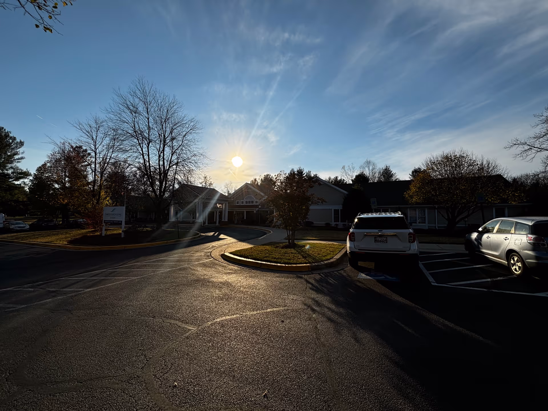 Entrance and driveway of a senior living building at sunset with parked cars and trees.
