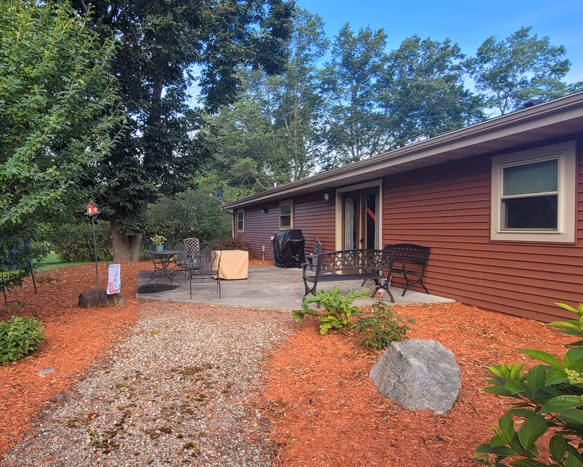 Patio area beside a single-story red-sided building with outdoor furniture, mulch beds, trees and a gravel path.