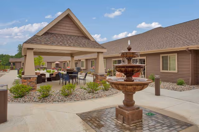 Outdoor courtyard of a senior living facility featuring a three-tier fountain, covered seating area, patio furniture, and single-story brown buildings.