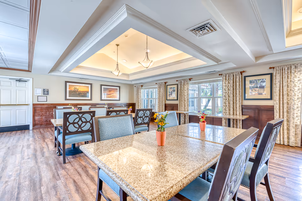 Bright communal dining room with granite-top tables, upholstered chairs, pendant lighting, and small flower centerpieces by large windows.