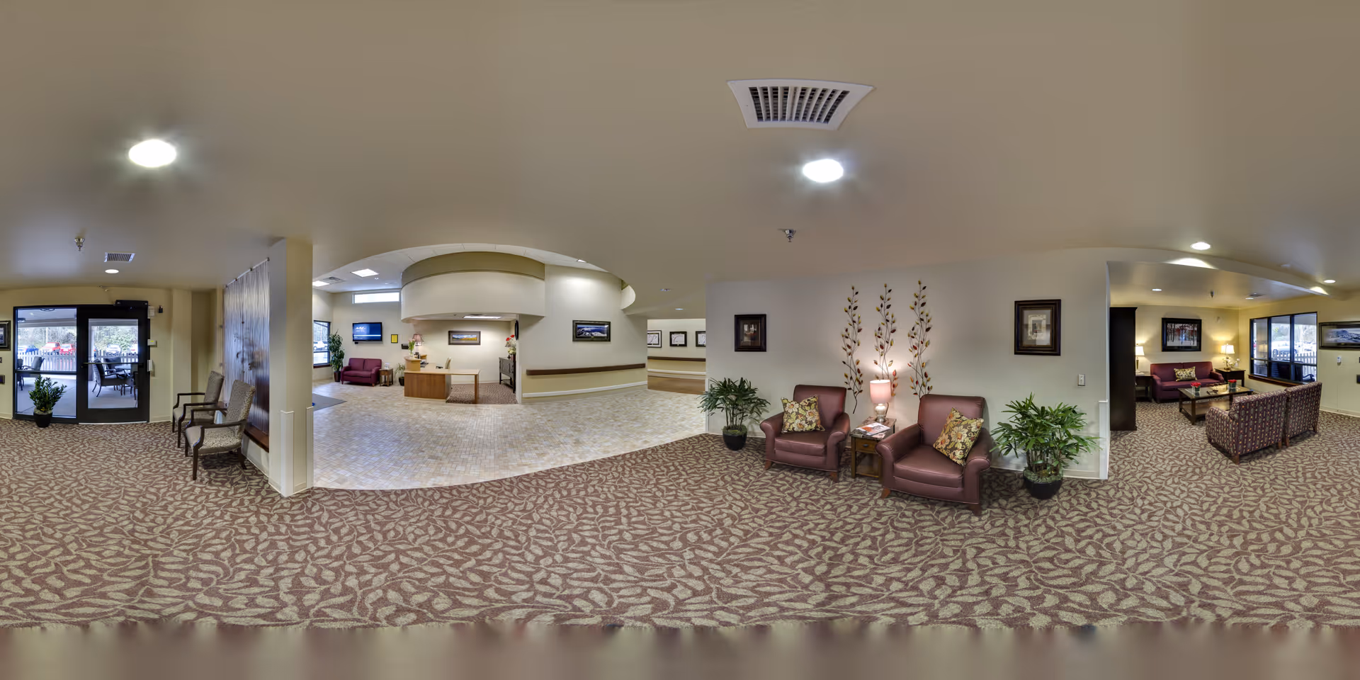 A spacious senior living facility common area with patterned carpet flooring and beige walls. The room features multiple seating areas with armchairs and sofas in shades of maroon and purple, side tables with lamps, potted plants, and framed artwork on the walls. There is a reception desk in the center background and glass doors leading to an outdoor patio area on the left side.