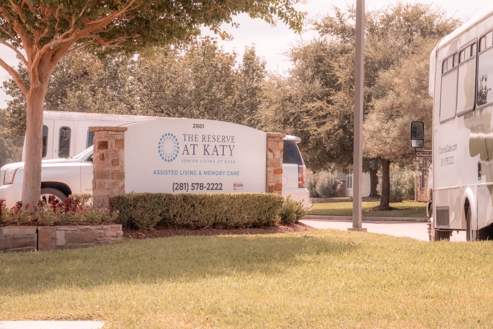 Outdoor view of the entrance sign for The Reserve at Katy Assisted Living and Memory Care, surrounded by greenery and trees, with vehicles parked nearby.