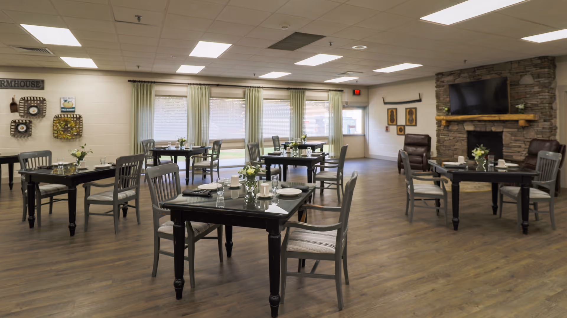 A spacious dining room with several black wooden tables and gray chairs arranged neatly. Each table is set with plates, glasses, and small flower vases. The room features large windows with green curtains allowing natural light to enter. There is a stone fireplace with a mounted TV above it and two brown leather chairs nearby. The floor is wooden, and the ceiling has recessed lighting panels.