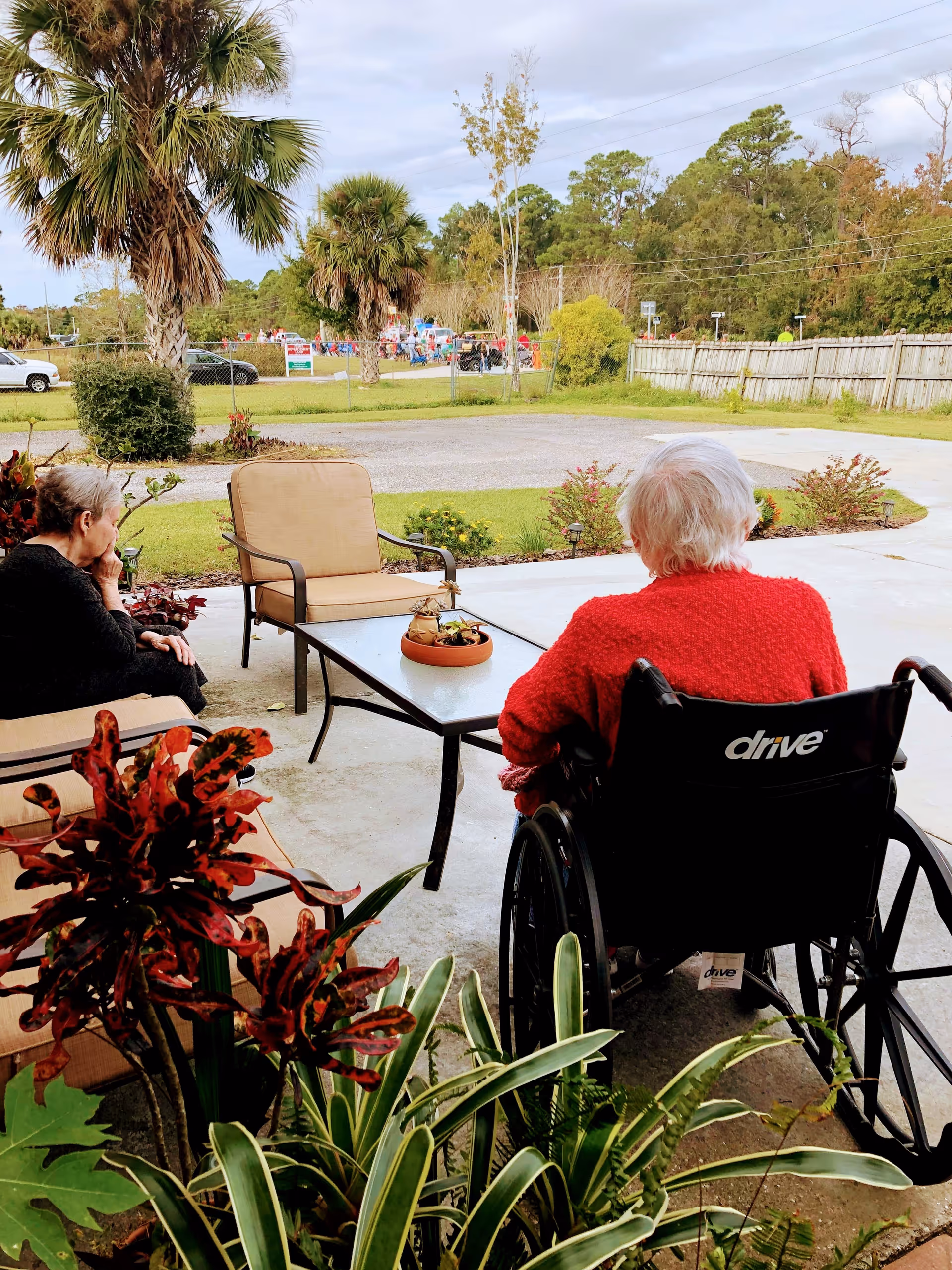 Two elderly individuals sitting outside on a patio area. One person is seated in a wheelchair wearing a red sweater, and the other is sitting on a cushioned chair. There is a glass-top table with a small potted plant between them. The background shows a grassy area with palm trees, a fence, and a parking lot with cars.