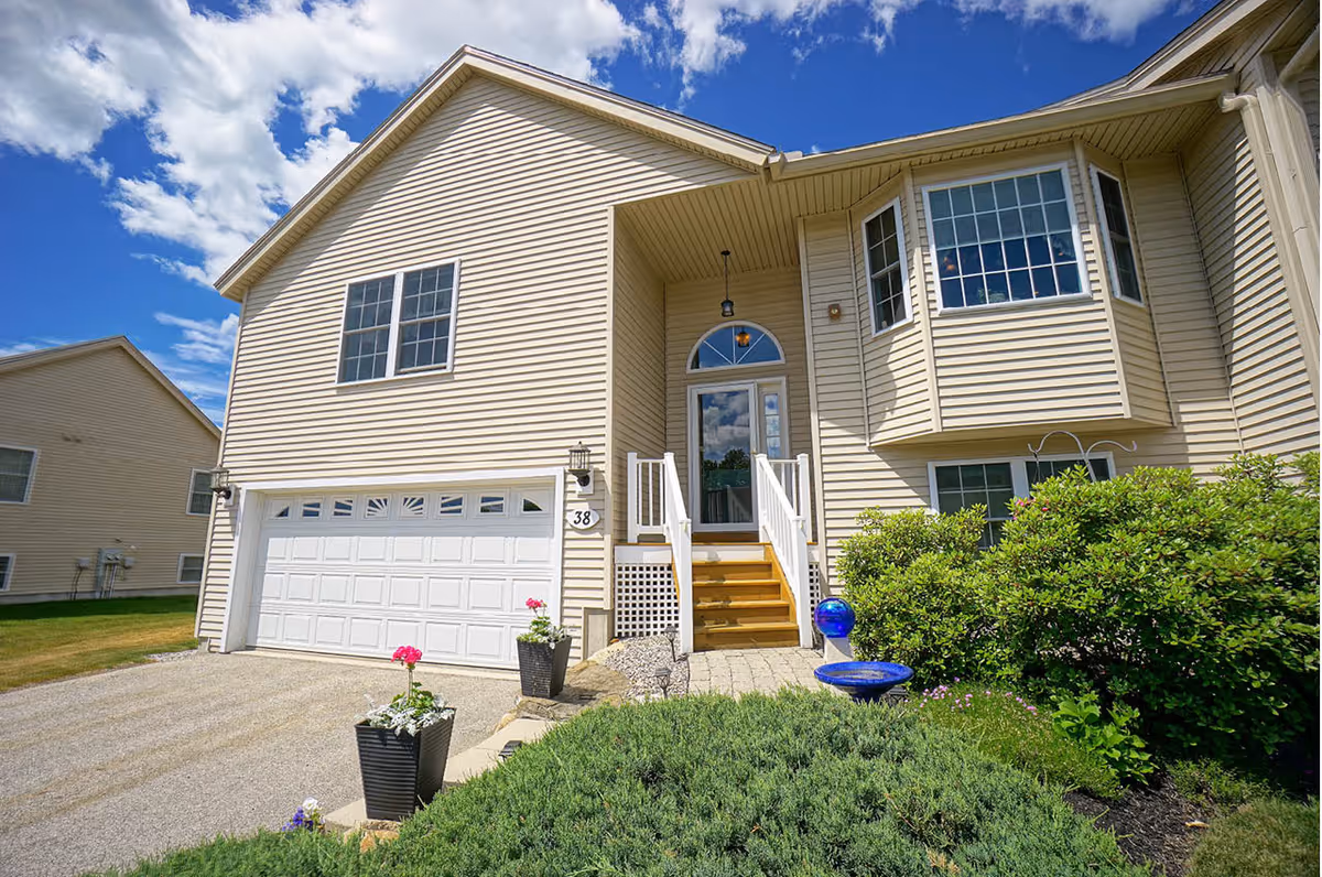 Exterior view of a beige two-story house with a white garage door, a small staircase leading to the front door, and a garden with green shrubs and potted flowers under a partly cloudy blue sky.