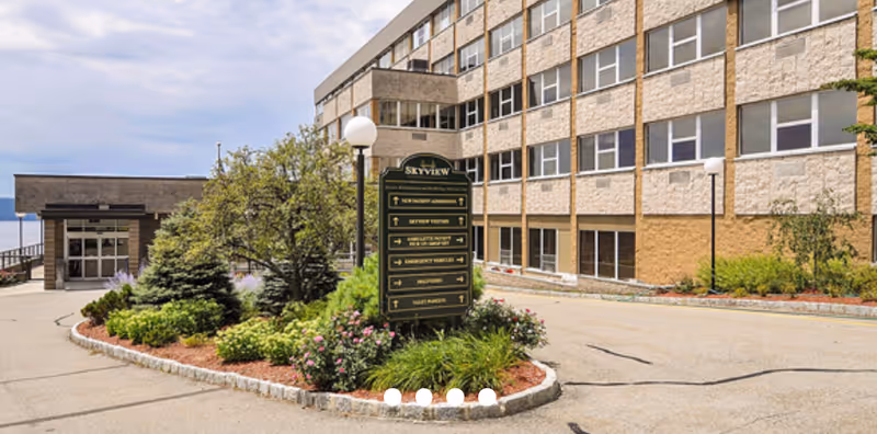 Exterior view of Sky View Rehabilitation & Health Care building with a multi-story structure, a landscaped area with bushes and flowers, and a signpost indicating directions to various facility areas.