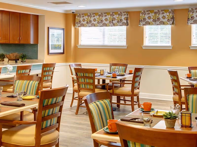 A bright dining room with wooden tables and chairs featuring striped cushions. The tables are set with orange cups, plates, glasses, and napkins. The room has light yellow walls, three windows with floral valances, and a kitchen area with wooden cabinets in the background.