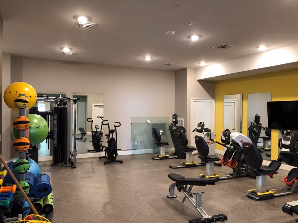 Indoor fitness room with exercise bikes, weight bench, and various exercise balls and mats stacked on a rack. The room has mirrors on the walls and a yellow accent wall with a mounted TV.