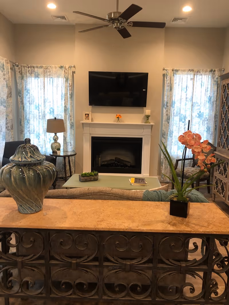 A cozy living room with a ceiling fan, two windows with floral curtains, a wall-mounted flat screen TV above a white fireplace, and a coffee table with decorative items. There are chairs on either side of the fireplace and a decorative cabinet on the right. In the foreground, there is a table with a large blue ceramic vase and a small plant with orange flowers.
