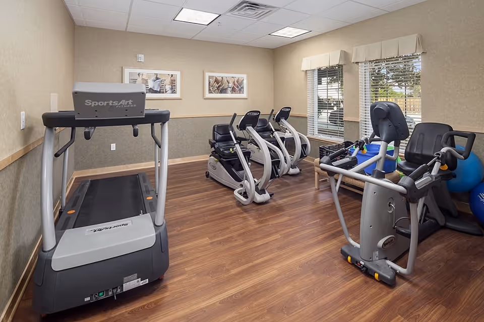 A small fitness room with exercise equipment including a treadmill, two recumbent stationary bikes, and another exercise machine. The room has wood flooring, beige walls with a chair rail, two framed pictures on the wall, and two windows with blinds and valances letting in natural light.