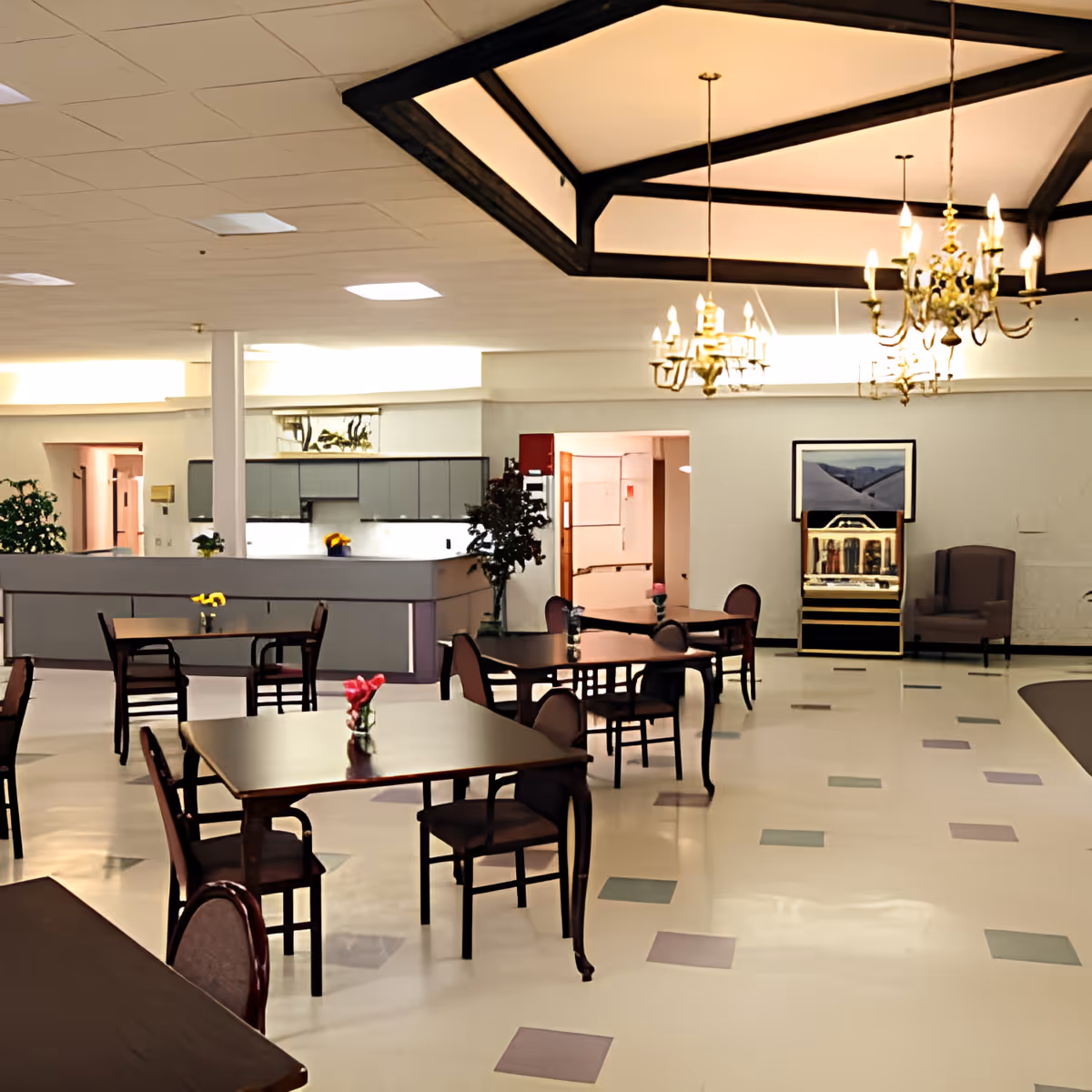 A spacious dining area with several dark wooden tables and chairs, each table decorated with a small vase of flowers. The room features a high ceiling with elegant chandeliers and a patterned floor with square tiles in various colors. In the background, there is a kitchen area with cabinets and a counter, as well as a jukebox and an armchair near the wall.