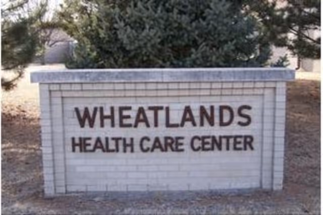 A brick sign reading "WHEATLANDS HEALTH CARE CENTER" standing outdoors with trees behind it.
