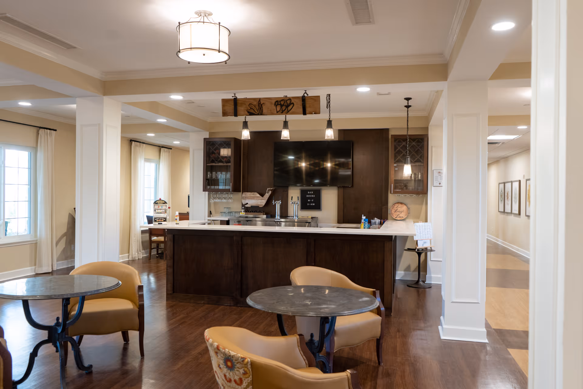 Interior view of a senior living facility lounge area with a dark wood bar counter, a mounted flat-screen TV, pendant lights, and small round tables with cushioned chairs. The room has large windows with white curtains, wooden flooring, and a hallway with framed artwork on the walls.