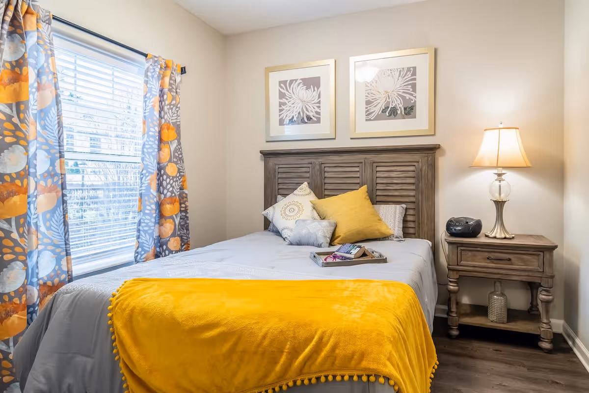 A cozy bedroom with a wooden headboard bed dressed in light gray bedding and a bright yellow throw blanket. There are three decorative pillows on the bed in white, gray, and yellow colors. A wooden nightstand beside the bed holds a lamp with a beige shade and a small black radio. Two framed floral artworks hang on the wall above the headboard. A window with colorful curtains featuring orange, gray, and white patterns is on the left side of the room.