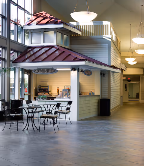 Indoor seating area with metal tables and chairs in front of a small food service counter labeled 'Beach Shack' inside a well-lit hallway with large windows and hanging ceiling lights.