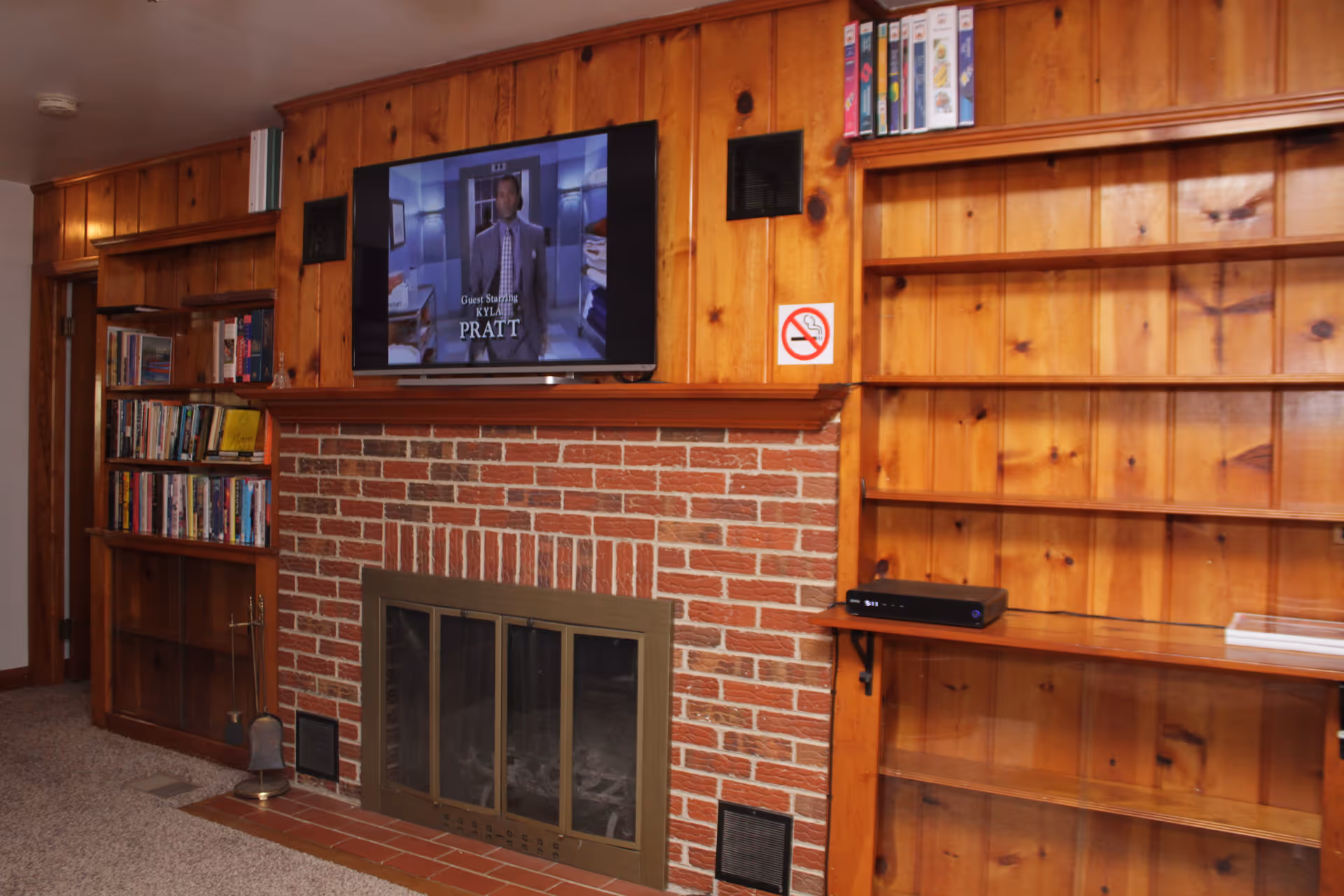 A cozy living room with wood-paneled walls and a brick fireplace with a metal screen. Above the fireplace is a mounted flat-screen TV displaying a show with the text 'Guest Starring KYLA PRATT'. On either side of the fireplace are built-in wooden bookshelves, one filled with books and the other mostly empty except for some items on the top shelf. A no smoking sign is mounted on the wall to the right of the fireplace.