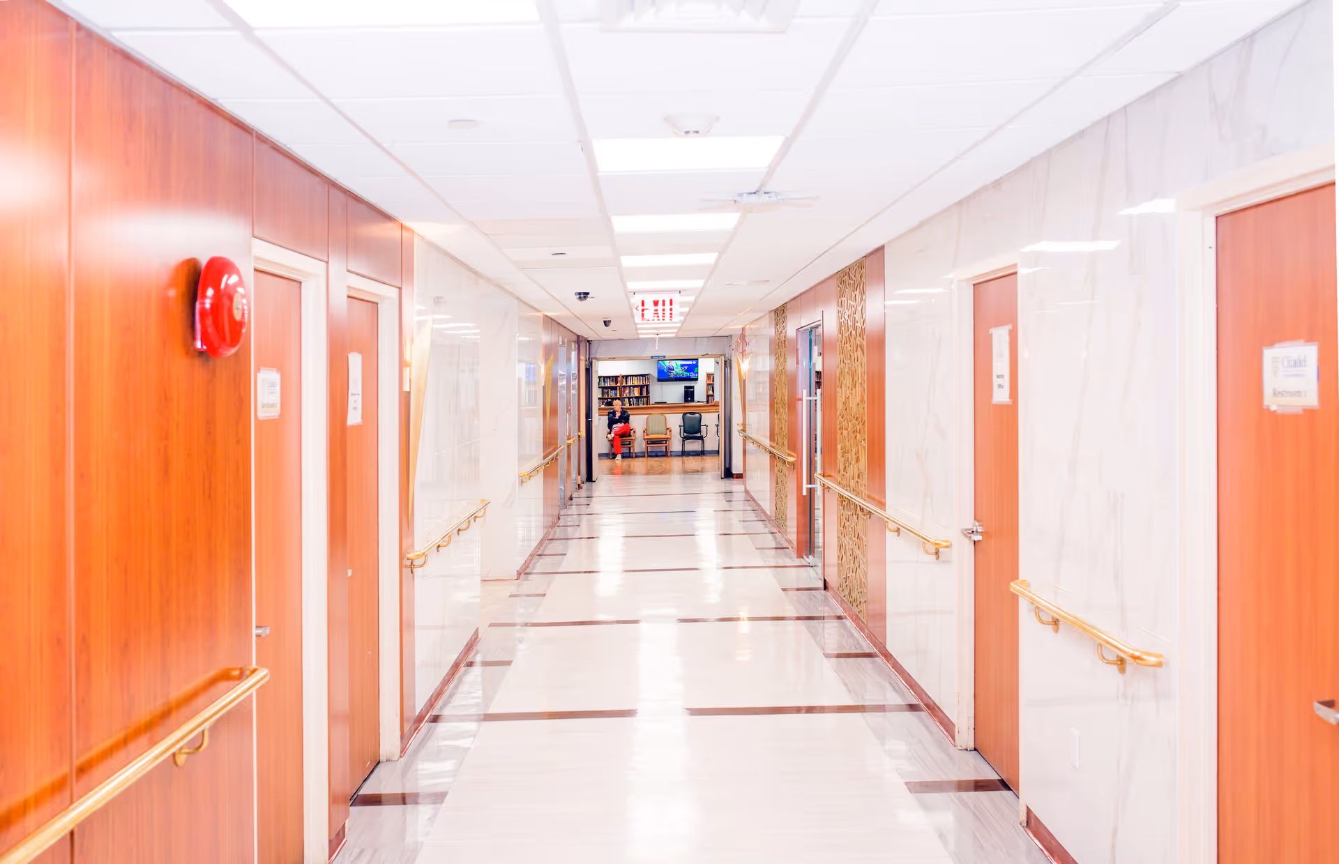 A brightly lit hallway in a rehabilitation and nursing center with wooden doors on both sides, handrails along the walls, a red fire alarm on the left wall, and a seating area with a person sitting at the far end under a television screen.