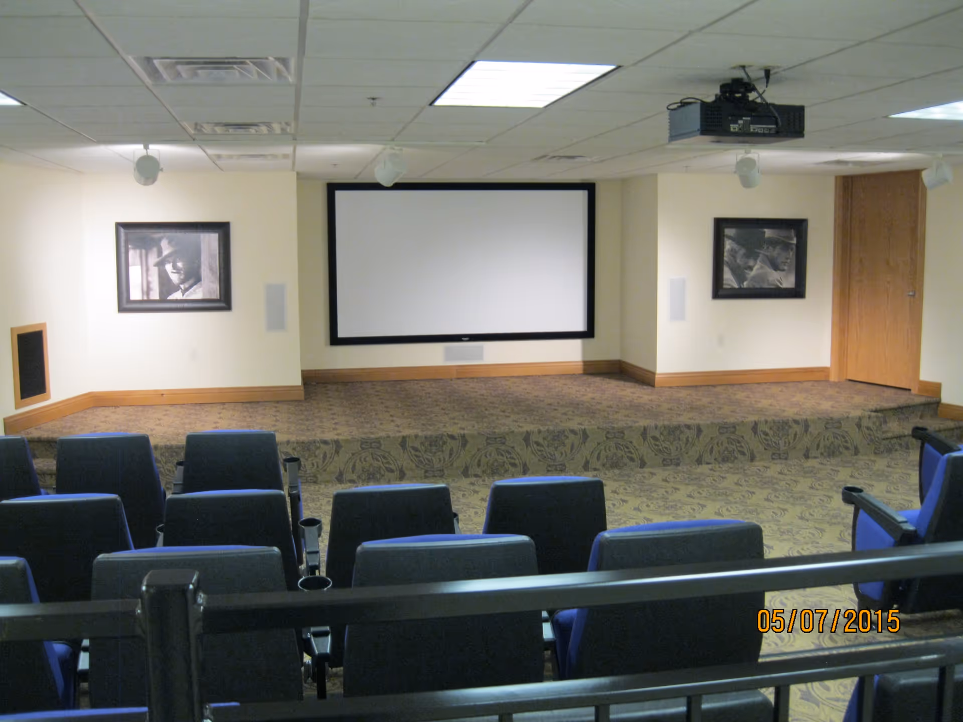 A small theater room with rows of blue cushioned seats facing a large blank projector screen mounted on a beige wall. The room has a patterned carpet and two framed black and white photographs on either side of the screen. A projector is mounted on the ceiling.