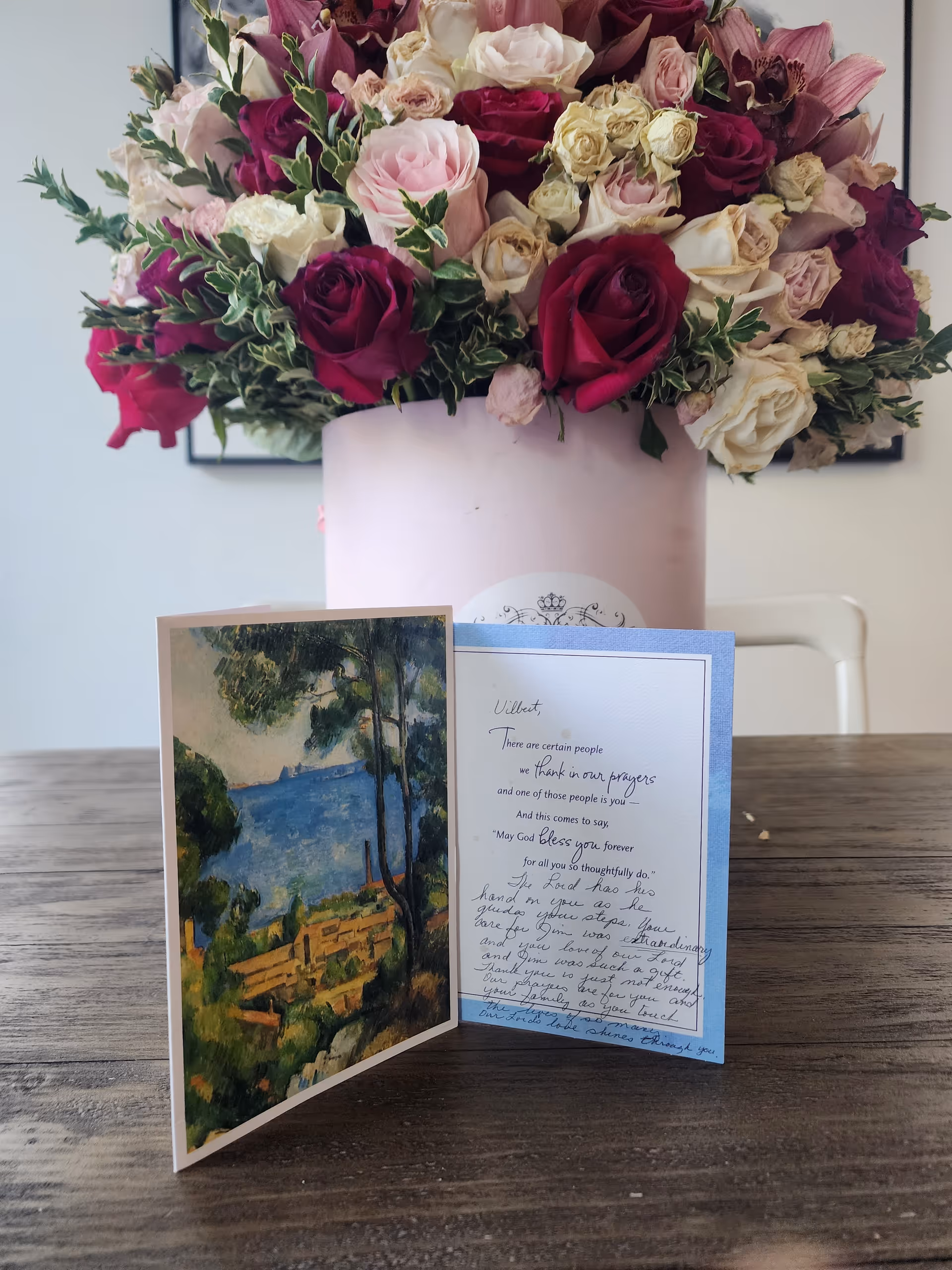 A large bouquet of mixed roses in shades of red, pink, and cream arranged in a pale pink cylindrical vase on a wooden table. In front of the vase, there is an open greeting card with a scenic painting on the left side and handwritten text on the right side. The background includes a white chair and a plain wall.