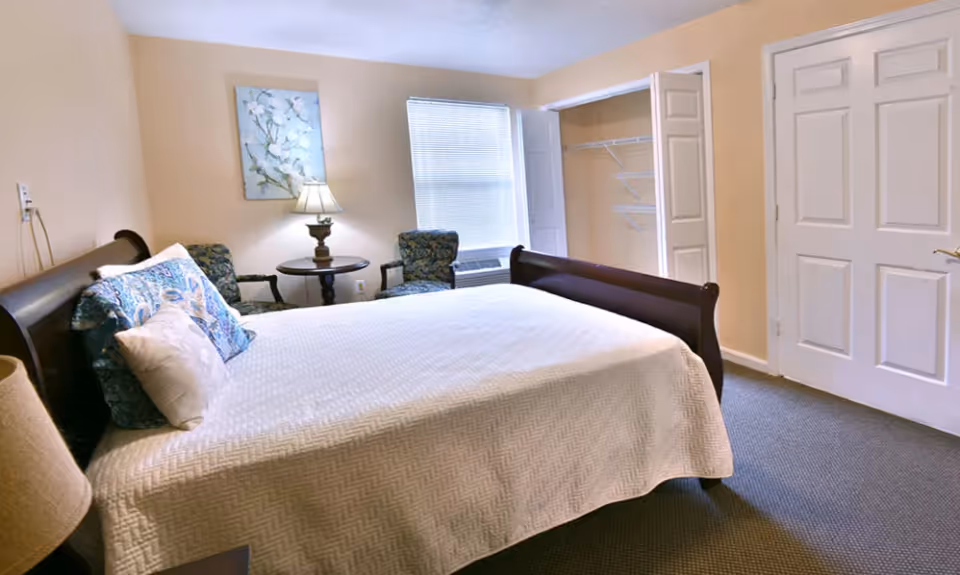 A bedroom with a large bed covered in a white quilt and multiple pillows. There are two upholstered chairs and a round wooden table with a lamp between them near a window with blinds. The room has beige walls, a carpeted floor, and an open closet with white doors.