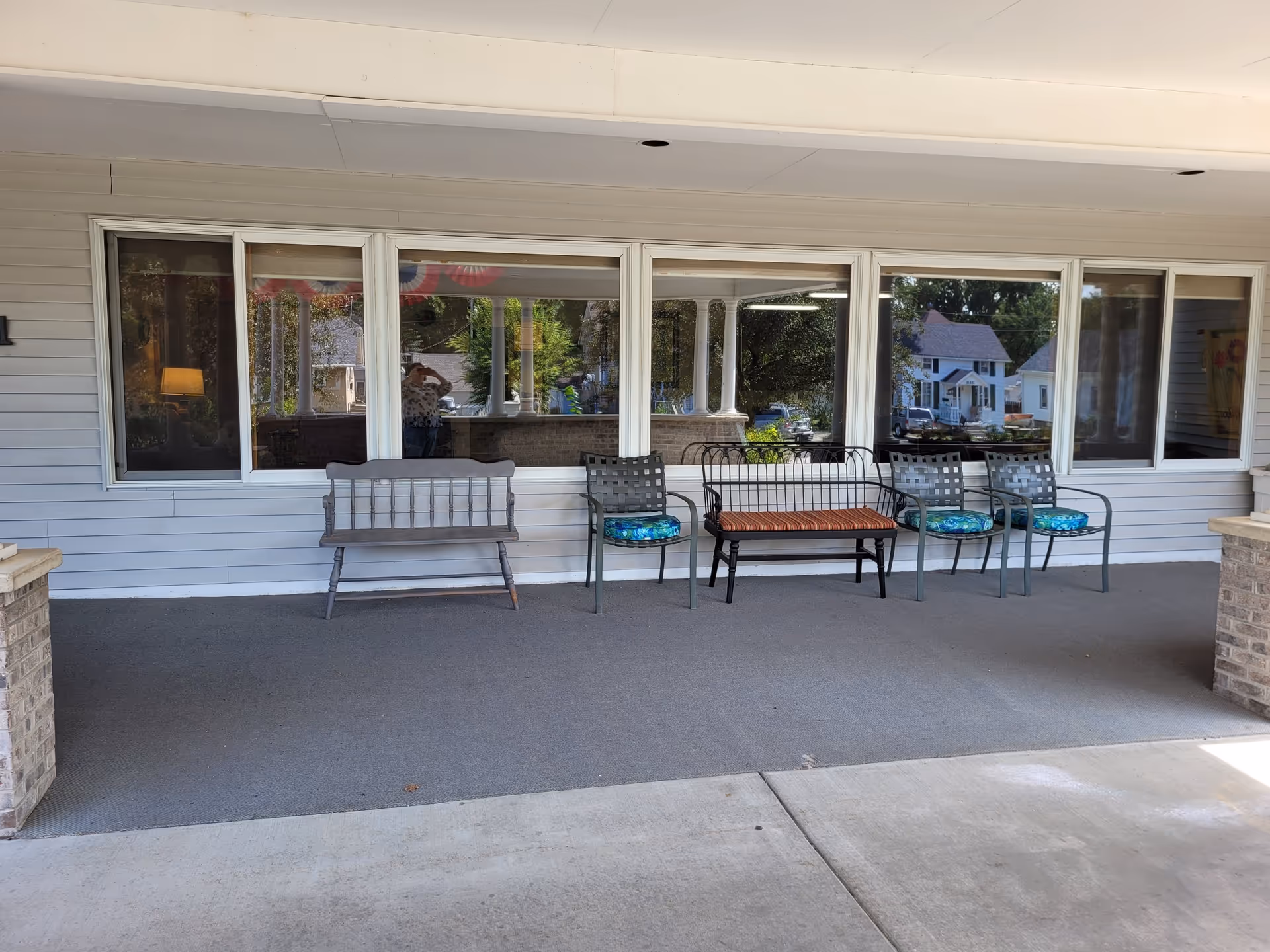 Covered outdoor seating area with a gray wooden bench, a black metal bench with a striped cushion, and three metal chairs with blue patterned cushions, in front of large windows reflecting trees and houses.