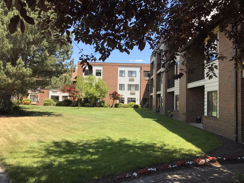 Outdoor view of King City Senior Village showing a well-maintained grassy courtyard surrounded by multi-story brick buildings with windows and balconies. Trees and shrubs are planted around the lawn, and the sky is clear and blue.