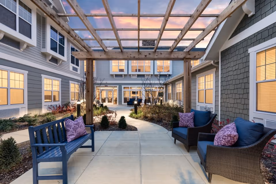 Outdoor courtyard area at The Commons on Meridian featuring a wooden pergola over a concrete pathway. There are benches and cushioned chairs with purple pillows on either side of the path, surrounded by landscaped plants and shrubs. The building exterior has multiple windows with warm interior lighting visible at dusk.