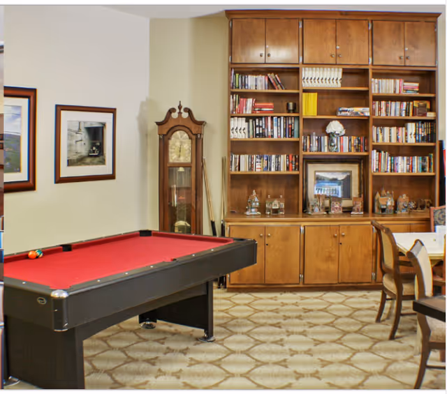 Pool table in recreation area with tall wooden bookshelf and chairs in senior living facility.