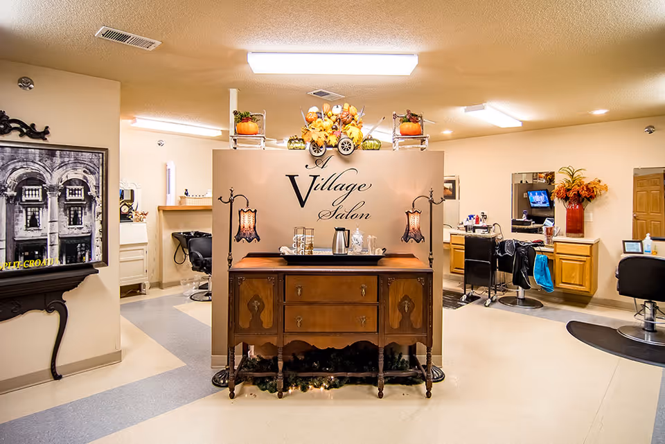 Interior view of a salon area named 'A Village Salon' with a wooden sideboard decorated with autumn-themed items and two vintage-style lamps. The salon has styling chairs, mirrors, and cabinets along the walls, with a framed black and white photo on the left wall and a vase with flowers on the right.