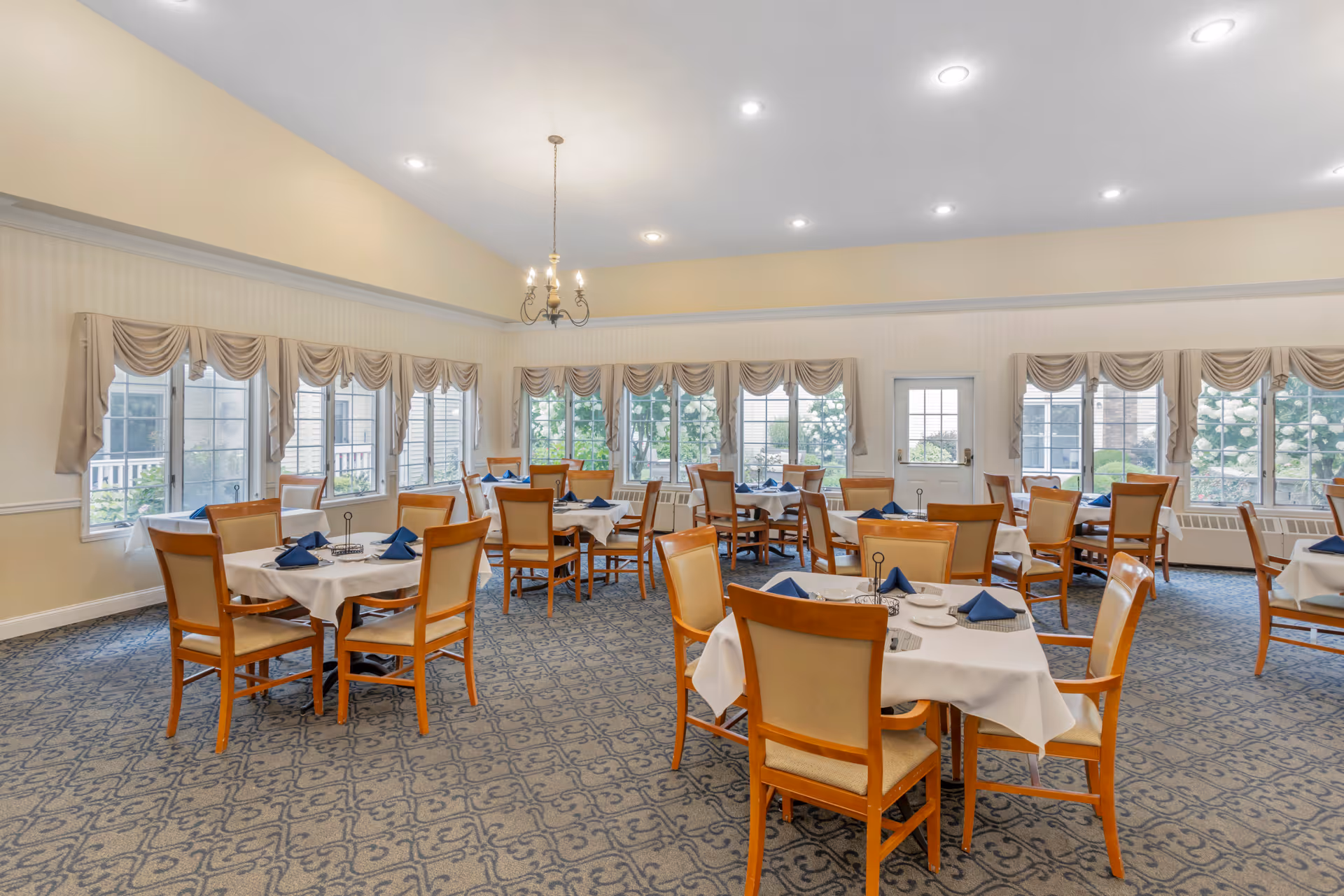 A bright dining room with multiple tables covered in white tablecloths and set with blue napkins. The room has large windows with beige curtains, a patterned carpet, and a chandelier hanging from the ceiling. Wooden chairs with beige cushions surround the tables.