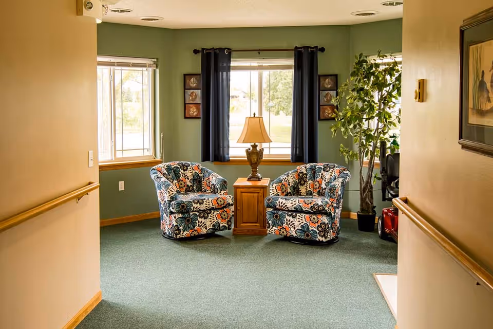 A cozy sitting area in a senior living facility with two floral-patterned armchairs facing each other, separated by a wooden side table with a decorative lamp. The room has green carpet, green walls with two windows covered by dark curtains, a potted plant in the corner, and framed artwork on the walls.