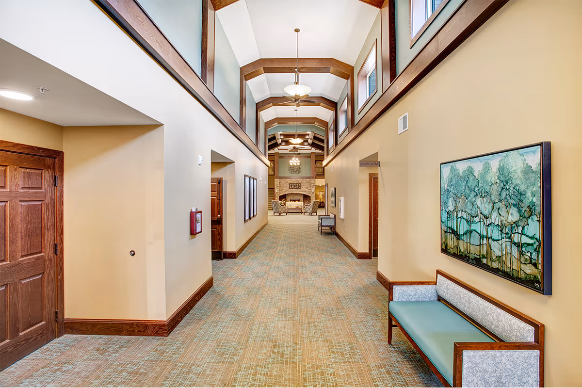 Wide carpeted hallway in a senior living facility with a high beamed ceiling, bench seating and artwork along the right wall leading toward a fireplace lounge.