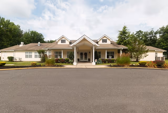 Single-story senior living facility facade with a central covered entrance, landscaping, and a paved driveway.