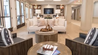 A spacious and well-lit living room area featuring a white sofa with beige cushions, two dark armchairs with patterned pillows, and two round wooden coffee tables. The room has large windows on the left side, a fireplace with a TV mounted above it on the far wall, and neutral-toned flooring and walls, creating a warm and inviting atmosphere.