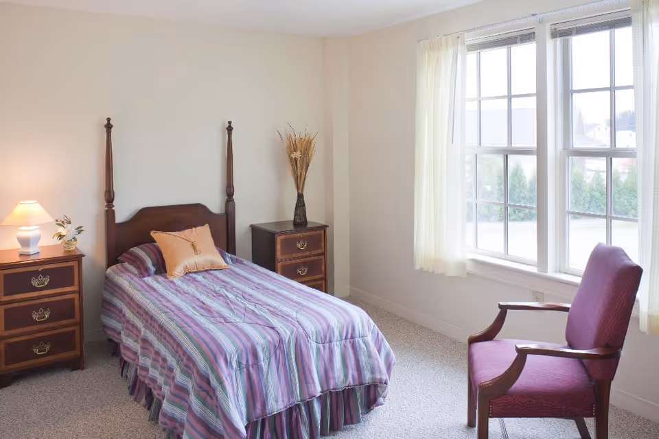 A simple bedroom with a single wooden bed covered with a striped purple and blue bedspread and a peach pillow. There are two wooden nightstands on either side of the bed, one with a lamp and a small plant, and the other with a tall vase holding dried plants. A purple upholstered chair is positioned near a large window with white curtains, letting in natural light.