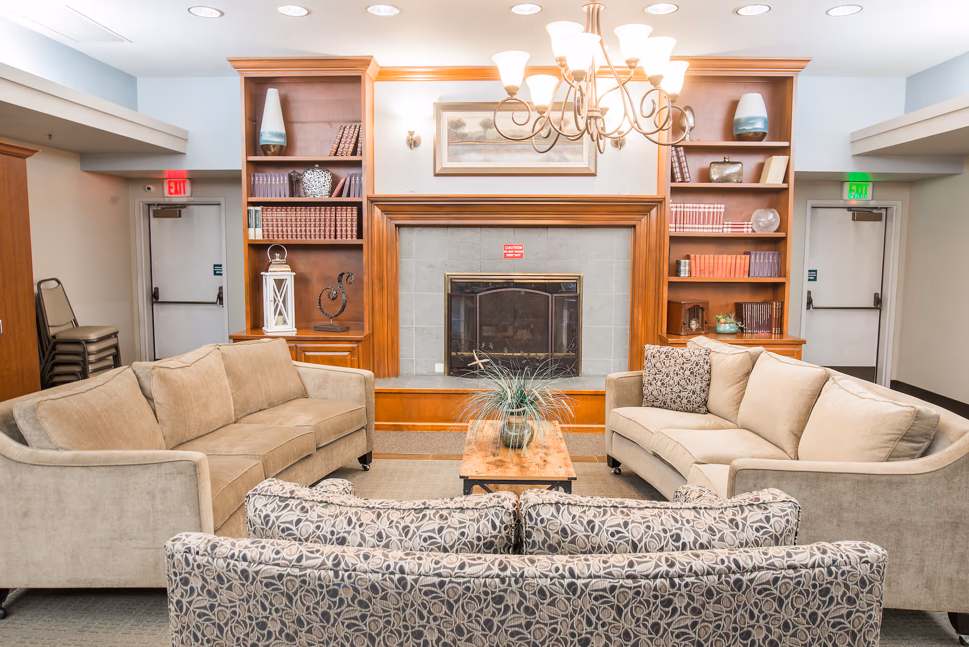 A cozy living room area with three sofas arranged around a wooden coffee table with a plant centerpiece. Behind the seating is a fireplace with a wooden mantel, flanked by built-in wooden bookshelves filled with books and decorative items. A chandelier hangs from the ceiling, and there are two exit doors on either side of the fireplace.