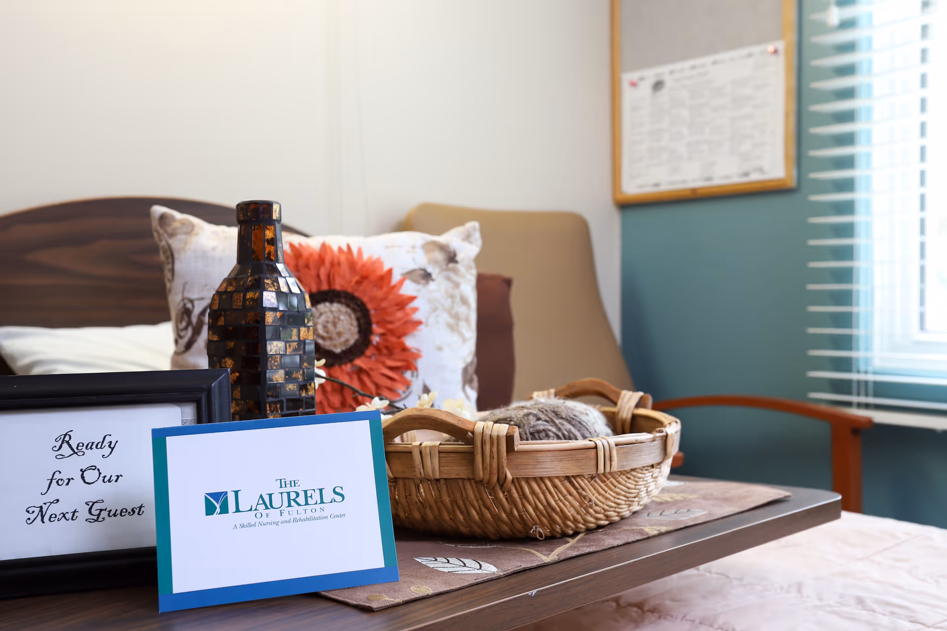 A decorated bedside table in a senior facility room with a wicker basket, decorative bottle, pillows, and a card reading 'The Laurels of Fulton'.