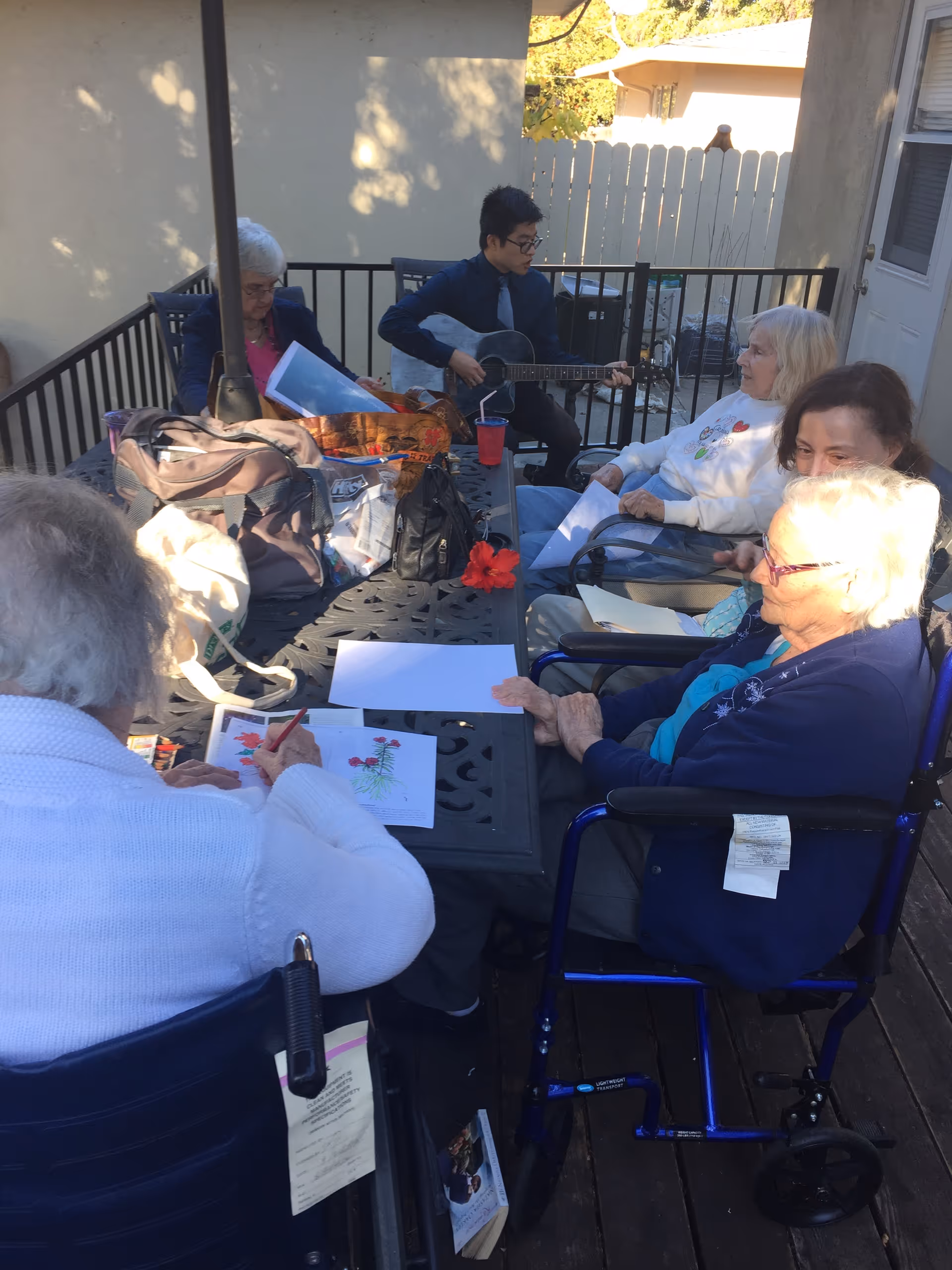 A group of elderly people sitting around a table on an outdoor patio. One person is playing a guitar while others are engaged in drawing or coloring activities. The setting is shaded with a fence and a building wall in the background.
