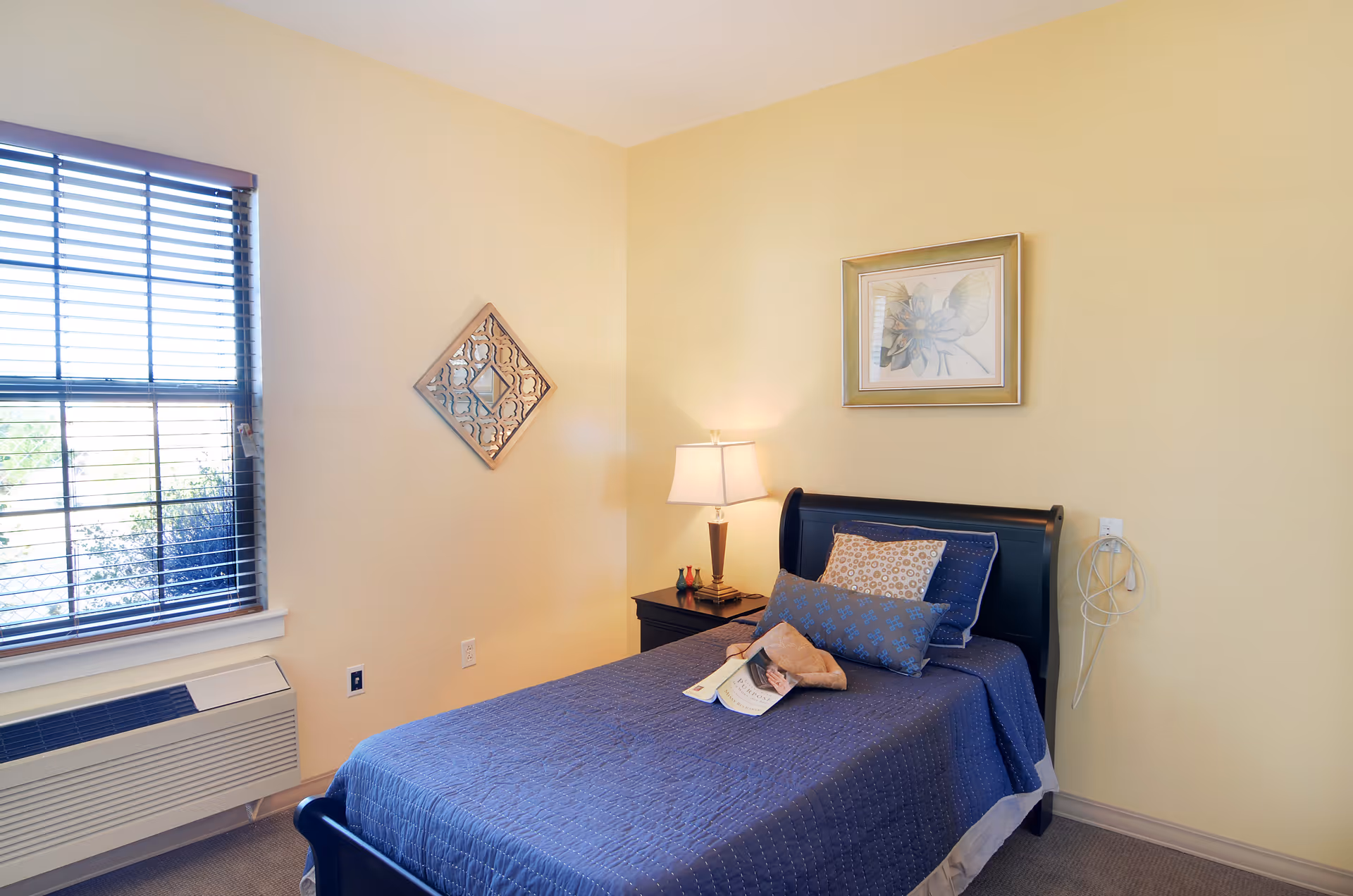A cozy bedroom with a single bed covered in a blue quilt and multiple pillows. There is a nightstand with a lamp and decorative items next to the bed. The walls are painted light yellow, with a framed floral picture above the bed and a decorative wall piece on the adjacent wall. A window with blinds allows natural light into the room, and an air conditioning unit is installed below the window.