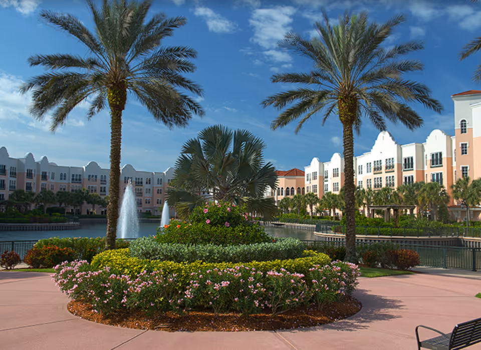 Landscaped courtyard with palm trees, a circular flower bed, fountains, and a multi-story building along a waterfront under a blue sky.