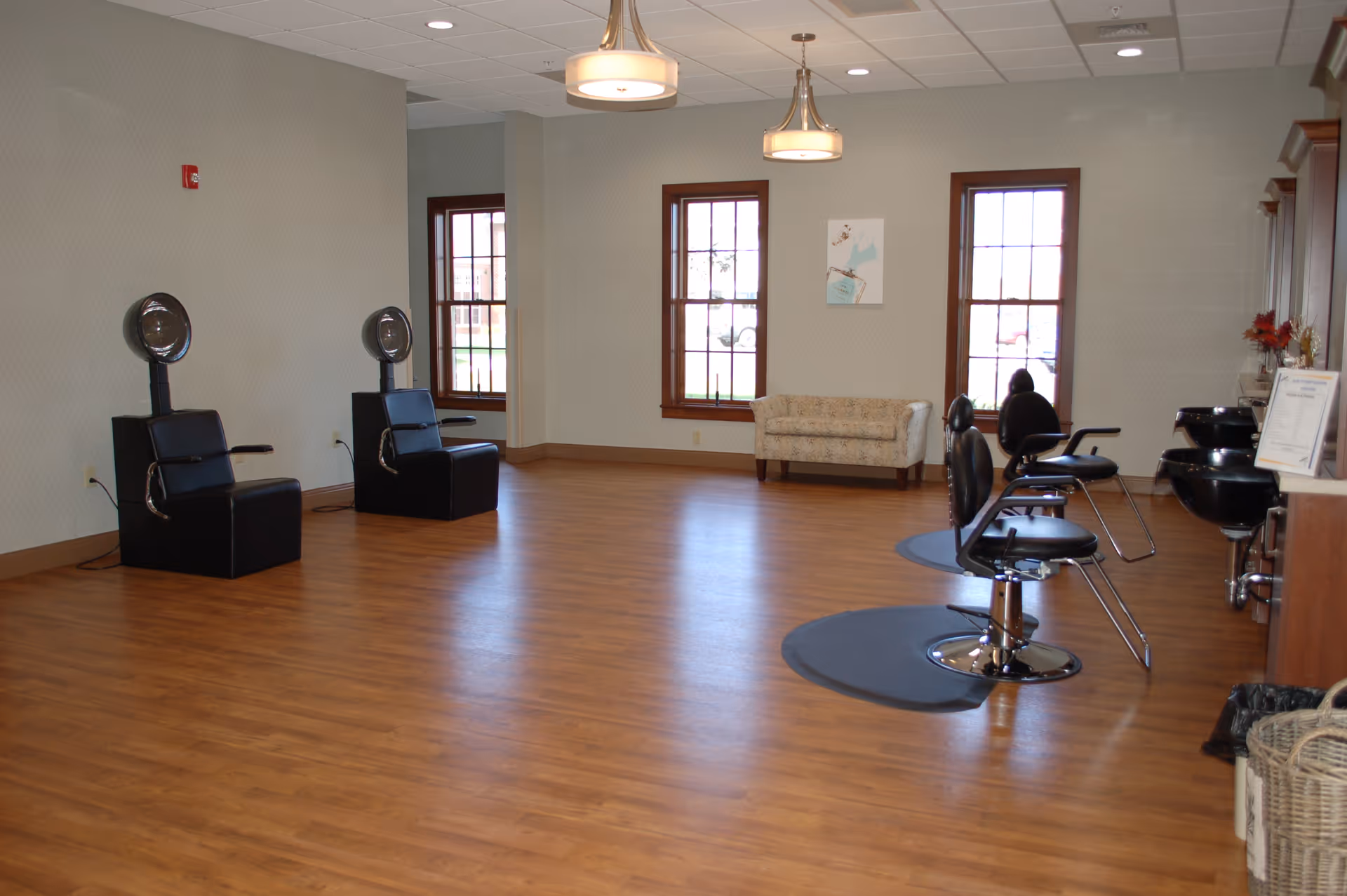 Interior view of a salon area in a senior living facility with wooden flooring, two black hair drying chairs on the left, three black salon chairs with sinks on the right, two large windows, a small beige couch, and two ceiling lights.