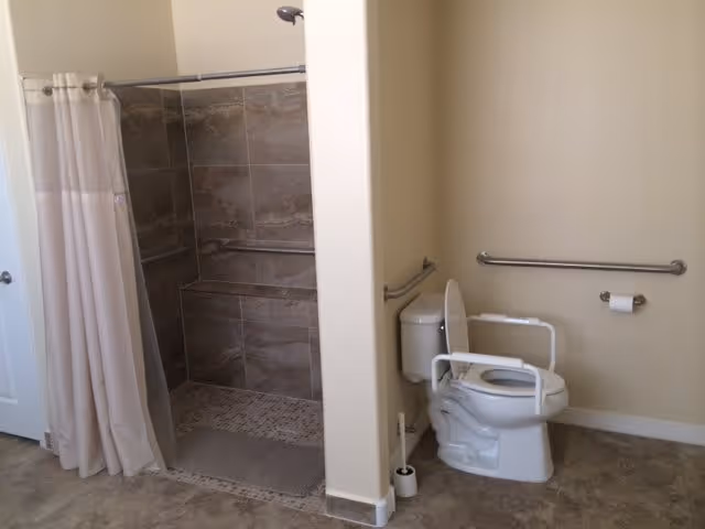 Accessible bathroom with a walk-in tiled shower featuring a built-in bench and a white shower curtain. Next to the shower is a toilet equipped with safety rails and grab bars on the walls for support. The floor is covered with beige tiles and the walls are painted a light cream color.
