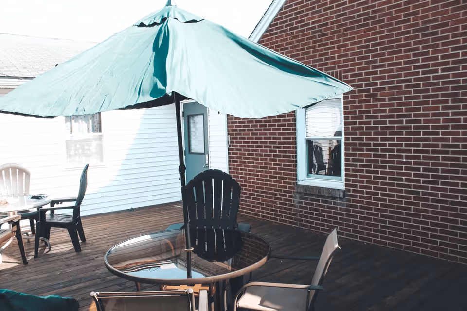 Outdoor wooden deck area with a round glass table, several chairs, and a large green patio umbrella. The deck is adjacent to a brick building and a white siding building with a door and window visible.