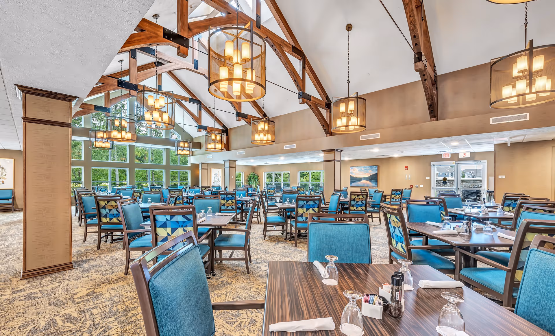 Spacious dining room in River Valley Retirement Community with wooden tables and blue cushioned chairs arranged neatly. Large windows allow natural light to fill the room, and multiple modern chandeliers hang from a high ceiling with exposed wooden beams.