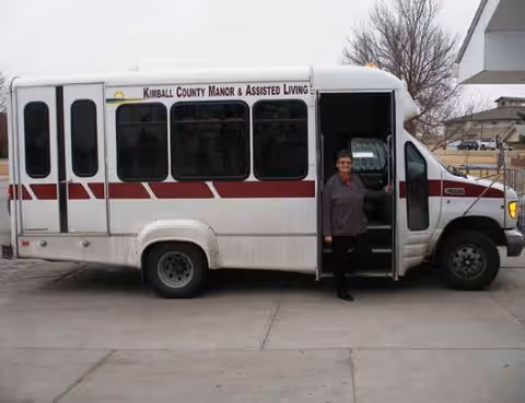 A white shuttle bus labeled Kimball County Manor; a person stands at its open door.