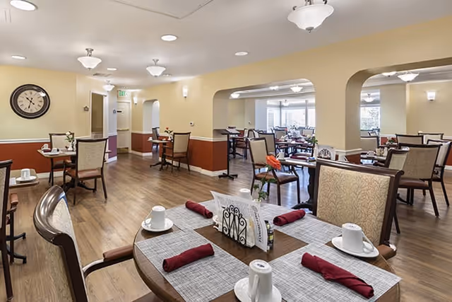 A spacious dining room in a senior living facility with multiple tables set with white cups, saucers, and red cloth napkins. The room features wooden flooring, beige walls with a red accent lower half, and several light fixtures on the ceiling. There are arched doorways leading to another dining area with large windows allowing natural light to enter.