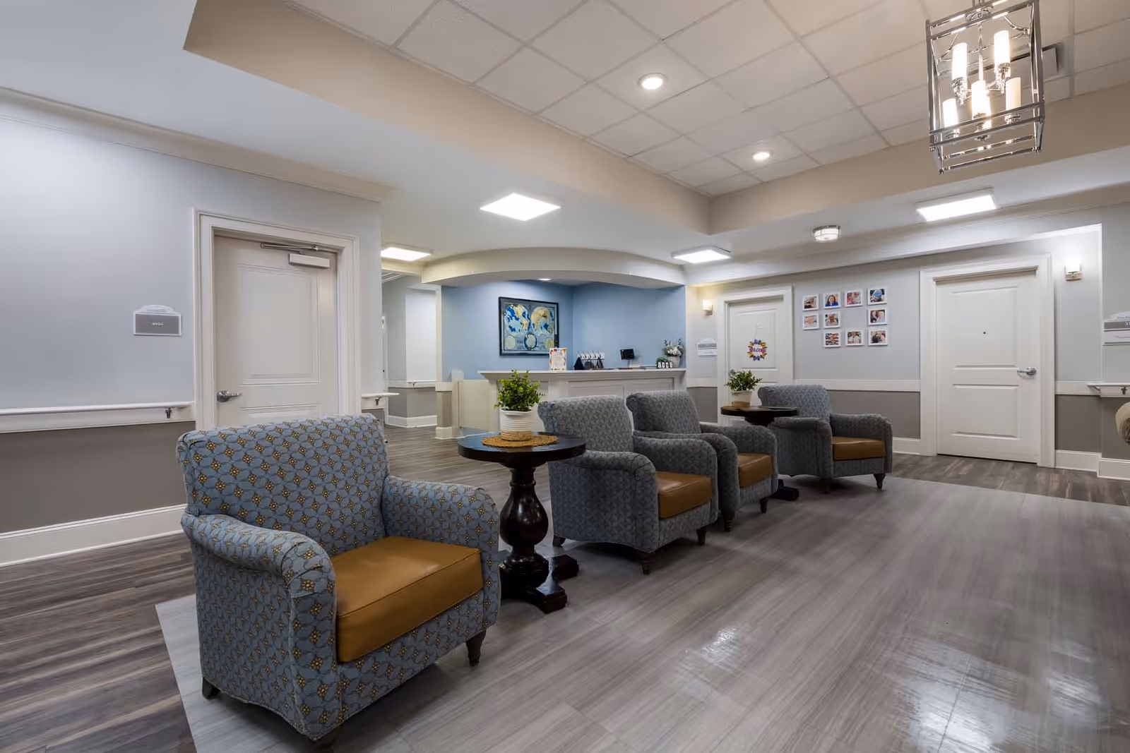 A bright senior living facility lobby with upholstered armchairs arranged around side tables and a reception desk in the background.