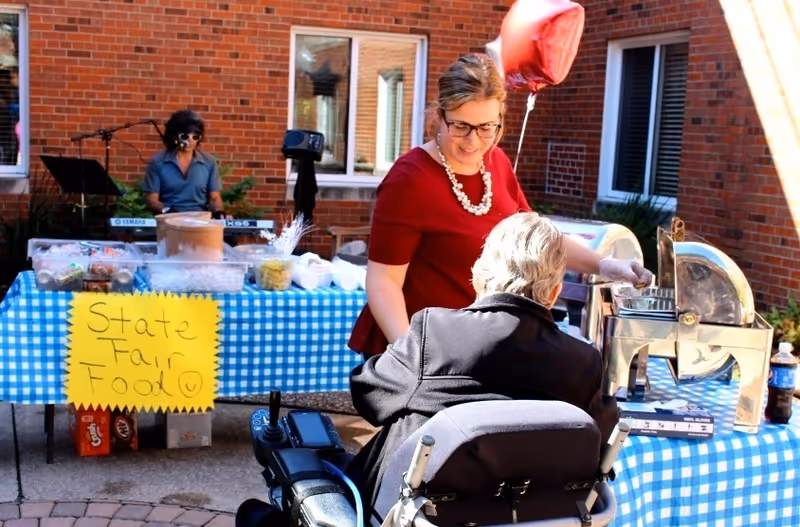 A woman serves food at a blue-checkered table labeled 'State Fair Food' to a person in a wheelchair in a brick courtyard.